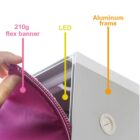 Close-up of a hand holding a 210g flex banner, partially exposing an LED light within an aluminum frame. Labels indicate each component: 210g flex banner, LED, and Aluminum frame.