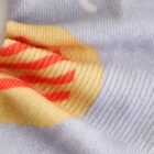 Close-up of a fabric featuring a vibrant abstract pattern with red and yellow stripes on a pale lavender background. The texture shows fine ribbing details.