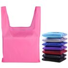 A bright pink reusable shopping bag is unfolded and upright. Next to it is a stack of six folded bags in varying colors: light blue, purple, navy blue, dark blue, black, and purple.