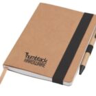 A brown notebook with a black elastic band and pen attached. The cover features the text Thumbtack Hardware. A bookmark ribbon is visible in the notebook.