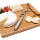 A bamboo cheese board with a round of Brie cheese, a cheese knife, crackers, and a small dish containing mixed olives, stuffed peppers, and feta cheese. The board has a carved wine bottle design on the surface.