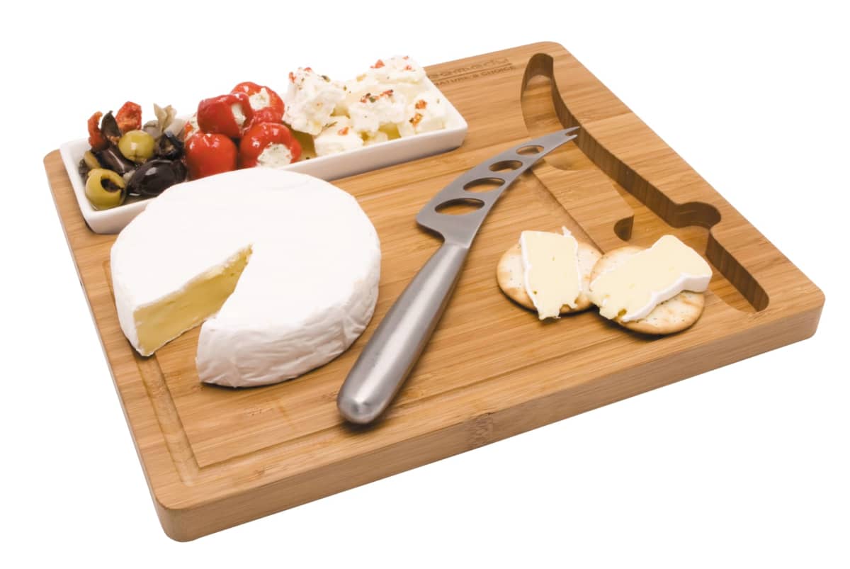 A bamboo cheese board with a round of Brie cheese, a cheese knife, crackers, and a small dish containing mixed olives, stuffed peppers, and feta cheese. The board has a carved wine bottle design on the surface.
