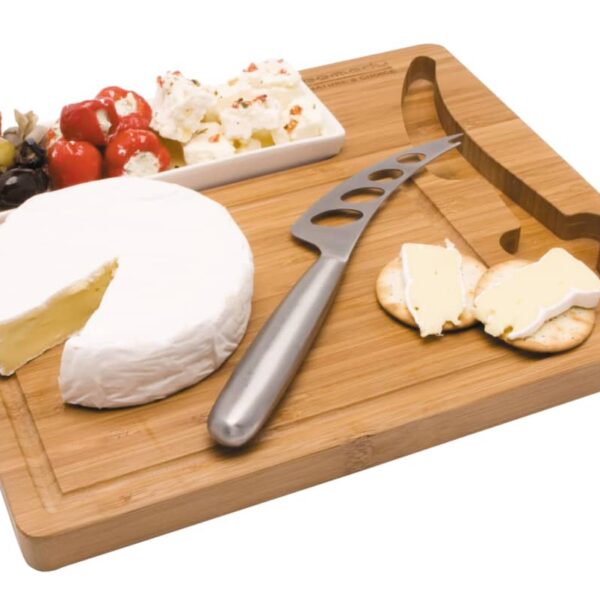 A bamboo cheese board with a round Brie, cheese knife, and crackers topped with cheese. A rectangular dish holds mixed olives and stuffed peppers.