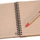 An open spiral-bound notebook with lined pages lies next to a pen with a beige body and red cap. The notebook features a recycling symbol in the bottom corners of each page.