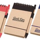 Three small notepads with spiral bindings and attached pens. Each has a different color scheme: red, black, and blue. The covers display the text Sink Em Swimwear.