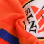 Close-up of an orange fabric with a partially visible white circular emblem featuring red triangles and a zigzag pattern. A blue stripe with dark edges is also visible on the fabric.