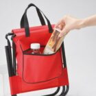 A red foldable chair with a built-in storage pocket. A persons hand is placing a carton into the pocket, which also contains a bottled drink. The chair has a black metal frame and black straps.