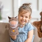 A young girl wearing a tiara and a blue dress is sitting on a couch, smiling and holding a zebra-striped hand puppet. A fuzzy brown puppet is partially visible on the right. The setting is a bright, cozy living room.