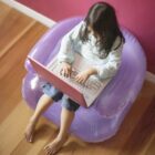 A young girl sits on a purple inflatable chair working on a pink laptop. Shes barefoot and wearing a light blue sweater with jeans. The floor is wooden, and the wall is pink.