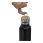 A hand lifts a metal loop attached to the wooden lid of a black stainless steel water bottle. The bottle is positioned upright with a plain white background.