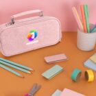 A pastel pink pencil case with a colorful logo is on a brown surface. Surrounding it are various stationery items, including pencils, colorful paper, sticky notes, clips, and rolls of tape. A pink wall is in the background.