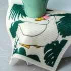 A green mug placed on a cloth with a white duck illustration featuring a yellow beak and feet. The duck is surrounded by green leaves, set on a marble surface.