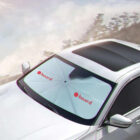 Car with a windshield sunshade featuring two red circles and the word board on each side. The sunshade is open, spanning the windshield. The car is parked, and there is a blurred background of trees and sky.