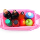 A top view of a pink bag containing various items: two bottles with corks, a dark bottle with a black cap, a blue carton, a red apple, a green apple, and a bottle with an orange liquid.