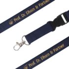 A navy blue lanyard with Prof. Dr. Dhom & Partner printed in gold lettering. The lanyard features a metal clip and a plastic buckle for easy attachment.