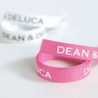 Two rolled ribbons are shown against a white background. One ribbon is pink with DEAN & DELUCA printed in white, while the other is white with the same text in black.