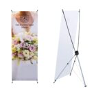 A floral-themed banner displaying a bouquet of pink and white flowers with the text The Flower Shop on the left. On the right is the backside of the banner stand, showing its frame and support structure.