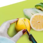 Hand with red glitter nail polish slicing a lemon on a bright green cutting board, next to a half lemon and avocado slices.