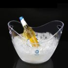 A transparent ice bucket filled with ice cubes holds a single bottle of beer with a blue bottle cap. The bucket has handles and sits against a dark background.