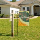 A For Sale sign stands in front of a suburban house. Next to it, a decorative flag reads Happy Thanksgiving and features autumn leaves and pumpkins. The house has a well-maintained lawn and a gable roof.