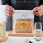 Person in an apron holding a clear bakery bag with a loaf of bread inside. Nearby are a sliced loaf on a cutting board and a glass with a butter knife on a napkin. The bag features a simple logo.