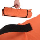 A hand is holding a rolled-up orange fabric bag with a black strap at the top. The bag features a black trim. There is a close-up of the strap securing the bag.