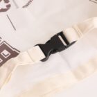 A close-up of a black plastic buckle fastened on a beige fabric strap, likely part of a bag or garment. The fabric has some printed text and graphics in brown visible around the buckle area.