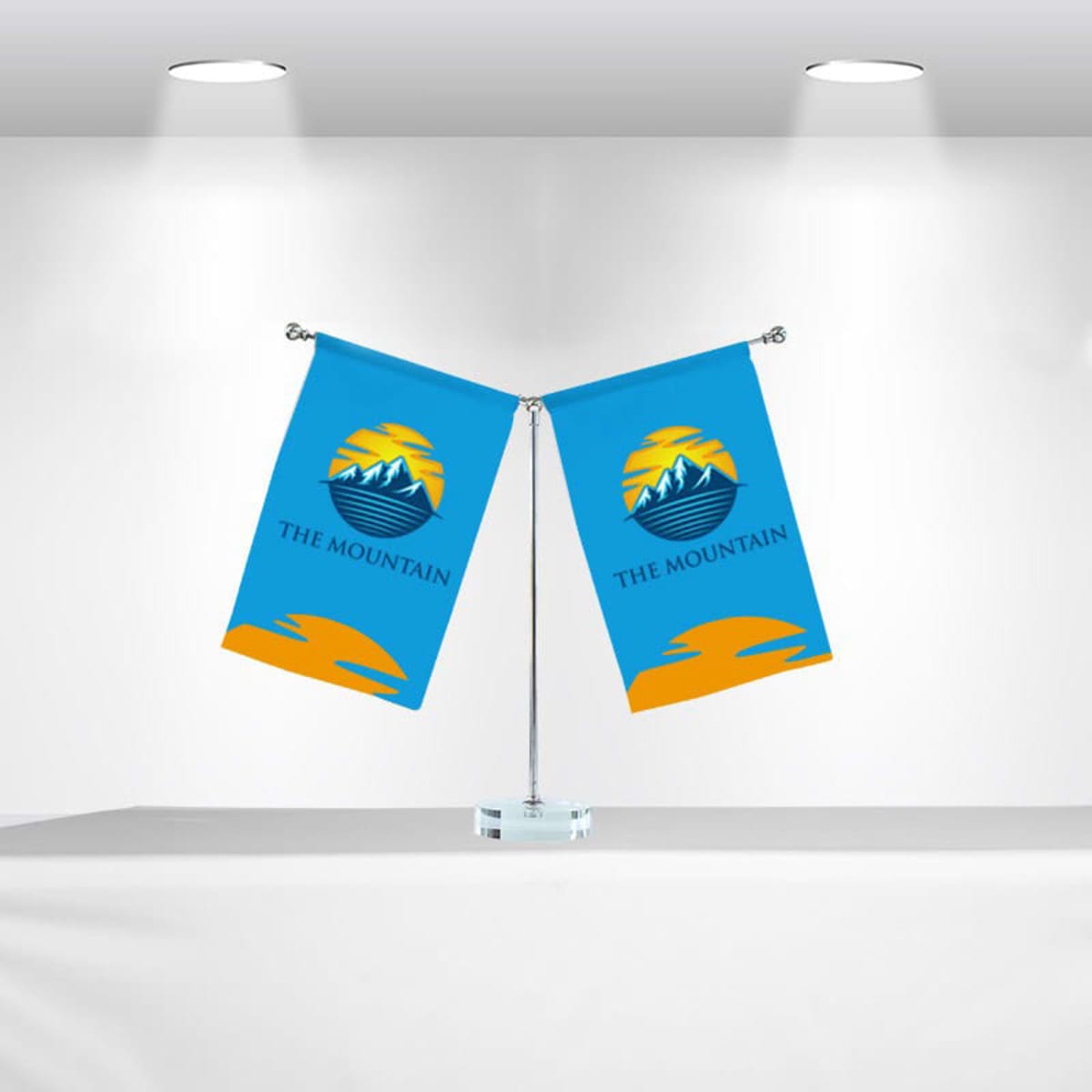Two blue flags with a mountain design and the words THE MOUNTAIN are displayed on a shared stand against a white background. Each flag features a stylized sun and mountain graphic in yellow and blue.