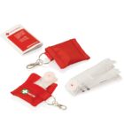 An image showing two red keychain pouches labeled First Aid, one closed and one open, revealing CPR masks. A white instructional booklet is displayed in the upper left corner.