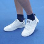 Close-up of a persons legs wearing white sneakers with white laces and navy blue socks, standing on a blue textured surface. The sneakers have a sleek design with a logo tag on the side and a pull tab on the heel.