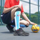 A person wearing a red soccer jersey, black shorts, and blue striped socks kneels on a sports court holding a yellow soccer ball. The court is fenced, with trees visible in the background under a clear sky.