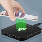 A hand holds a smartphone above a black wireless charging pad with a glowing green symbol. The pad is connected via a cable and is shown on a smooth, reflective surface.