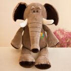 A plush elephant toy sits upright on a beige surface. It’s brown with large ears, an elongated trunk, and white tusks. The toy has a colorful logo with kids on its belly. Blurred pink flowers are in the background.