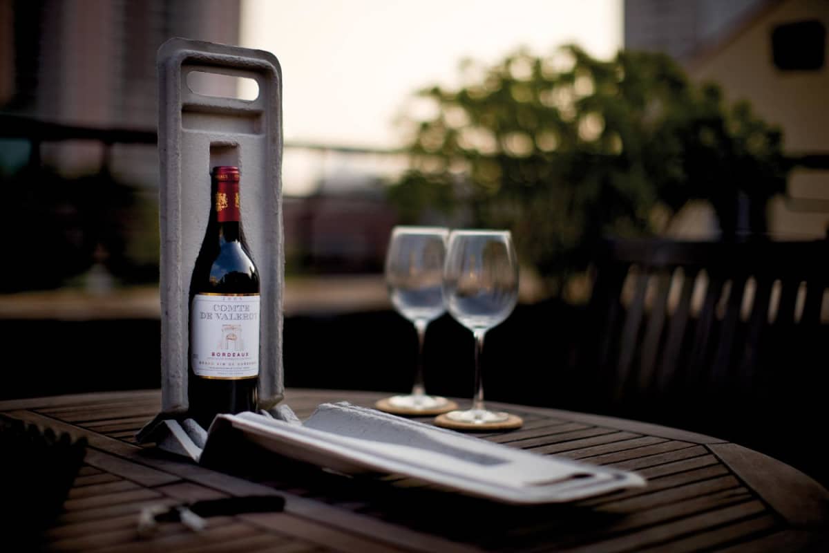 A bottle of wine in a holder on a wooden table, accompanied by two empty wine glasses. The setting appears to be an outdoor area with blurred buildings and greenery in the background.