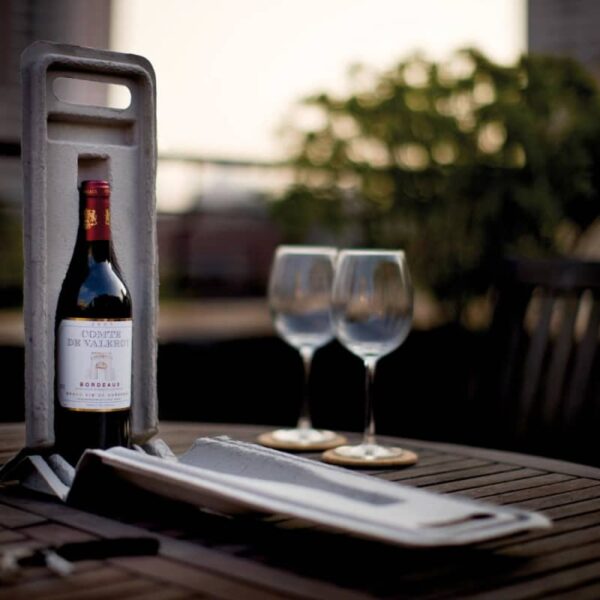 A bottle of red wine stands in a holder on a round wooden table, accompanied by two empty wine glasses. The background shows a blurred outdoor scene in soft focus, suggesting a patio or balcony setting.
