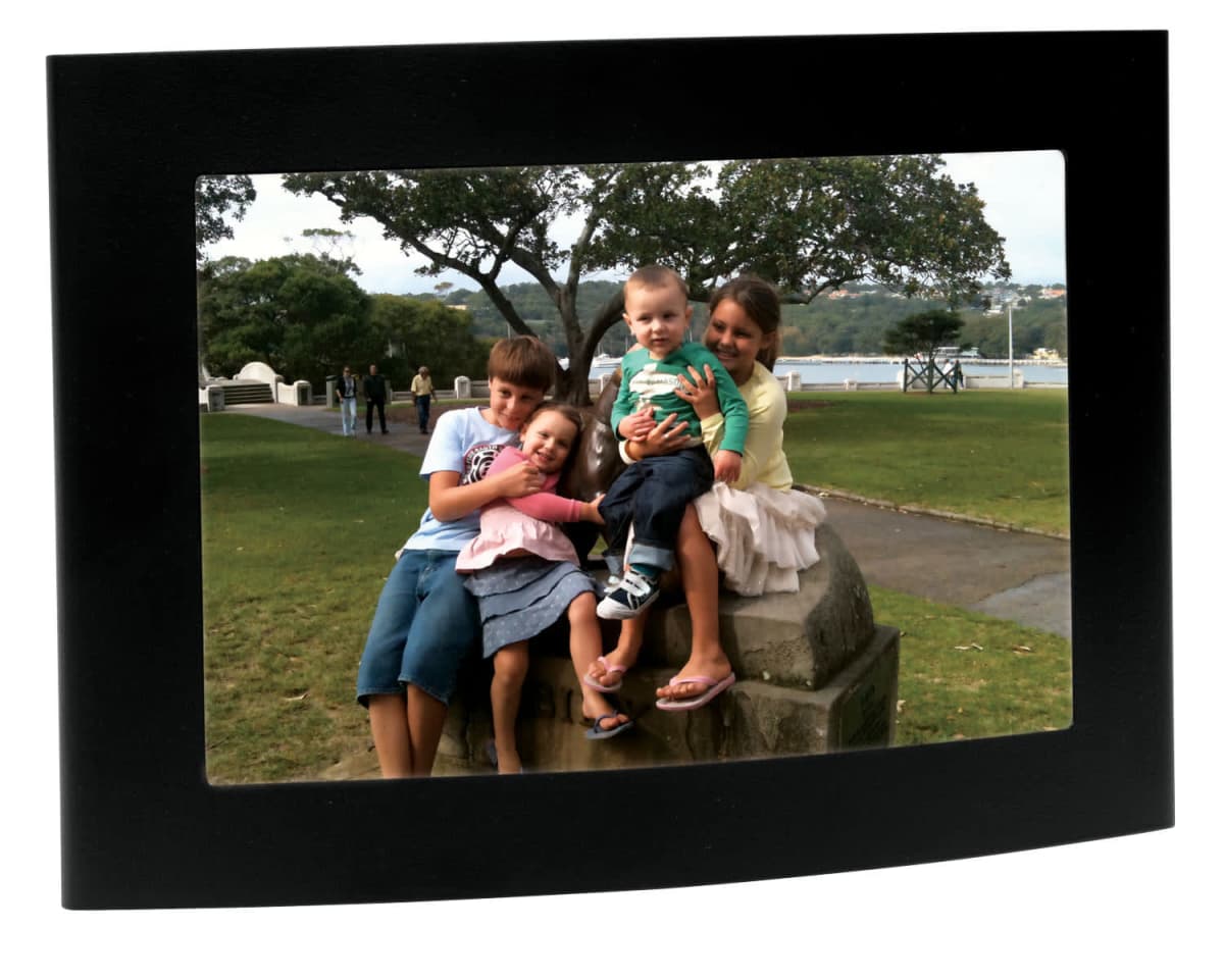 Four children sit together on a stone bench in a park, smiling for the camera. The background features trees, grass, and a view of the water. The image is displayed in a rectangular black frame.