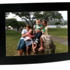 Four children sit on a stone bench outdoors near a body of water, with trees in the background. The group includes three older children smiling and holding a toddler. The scene is captured in a black, curved photo frame.