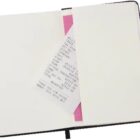 An open notebook with a pink ribbon bookmark lies flat. A printed receipt is partially tucked under the ribbon, with typed text visible on the receipt. The notebook has blank pages and a black cover.