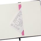 An open notebook with a pink sticky note and a receipt tucked inside, showing transaction details and a total amount of $12.00. The notebook is empty with white pages, and it has a black elastic band.