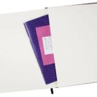 An open notebook with blank pages has a small pink folder and a purple folder tucked inside. A small handwritten note is visible in the pink folder pocket. The notebook has a black ribbon bookmark.