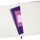An open notebook with blank pages, featuring a pocket. Inside the pocket, theres a purple and pink paper with white text partially visible. A narrow black ribbon serves as a bookmark. The notebook has a sleek and minimal design.
