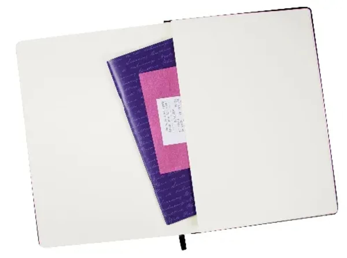 An open notebook with blank pages reveals a pocket containing a purple and pink booklet and a small piece of white paper. A black ribbon bookmark is visible at the bottom of the notebook.