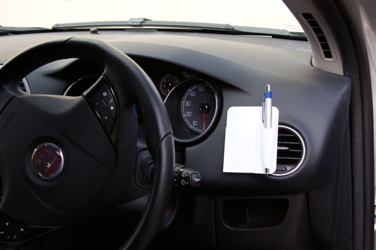Car interior showing the steering wheel, air vent, and dashboard. A small notepad with a pen is clipped to the air vent. The speedometer and other gauges are visible behind the steering wheel.