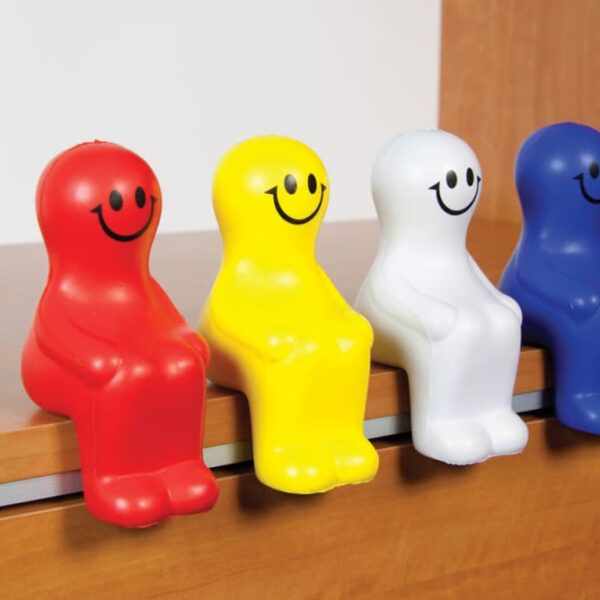 Four colorful smiling figurines sit on a wooden edge. From left to right, they are red, yellow, white, and blue. Each has a simple design with a round head, body, and two small legs.