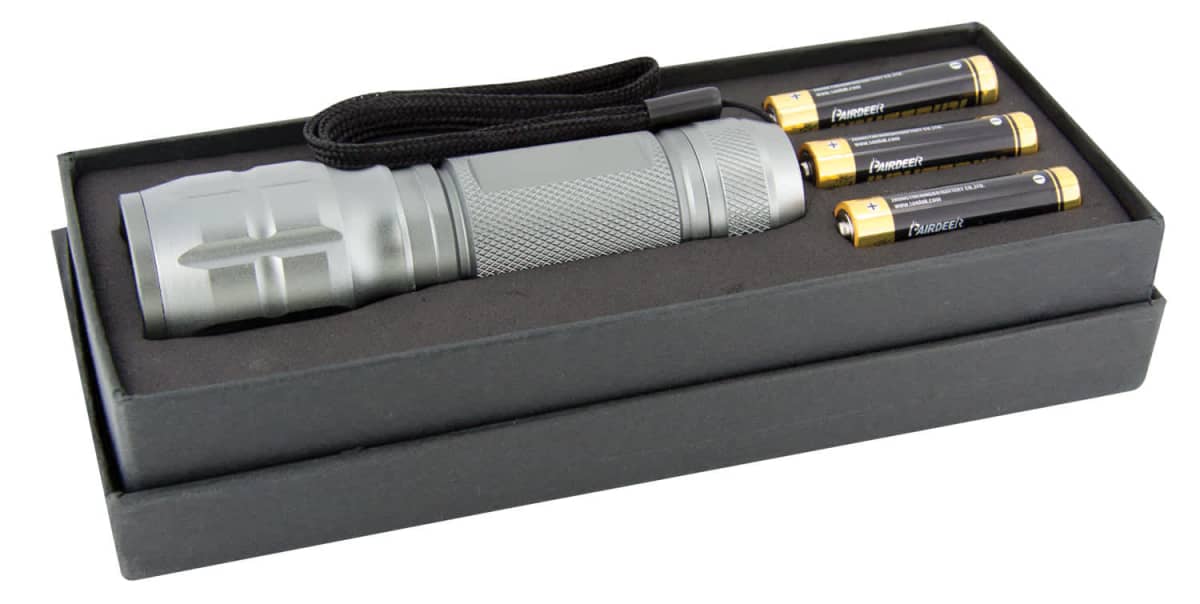 A silver flashlight with a textured grip and wrist strap is placed in an open black box. Three AA batteries are lined up next to it inside the box.