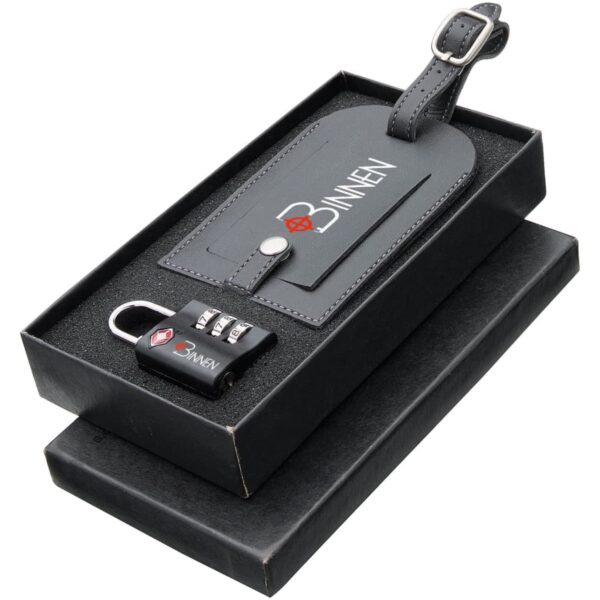 A black leather luggage tag with a long strap and a silver buckle is displayed in a black box. Next to the tag, there is a small black combination lock with three dials. Both items bear a logo with the letter B.