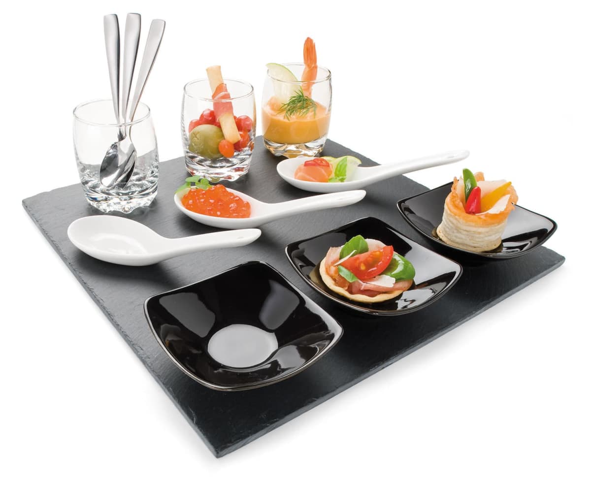 A slate platter with a sophisticated appetizer arrangement: three spoons with salmon and herbs, two glasses with seafood and dill, small black dishes with dipping sauce, and two canapés topped with tomatoes and greens.