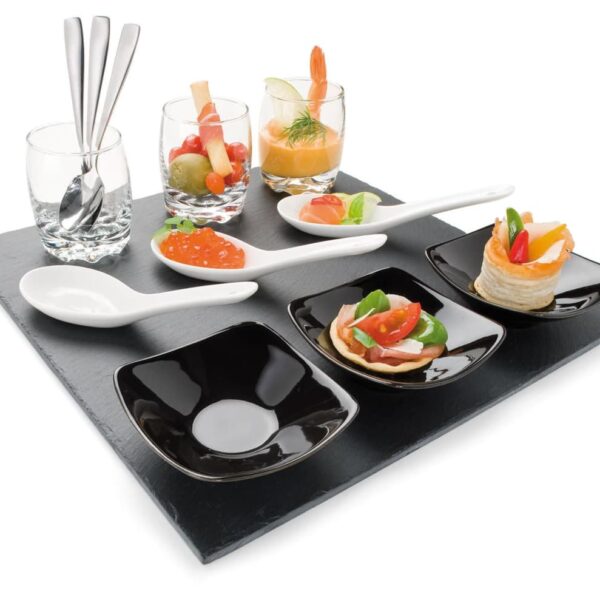 A slate board featuring an assortment of gourmet appetizers including spoons with salmon and dill, small glasses with shrimp and vegetables, and black dishes with bite-sized snacks. Three spoons rest in empty glasses on the left.