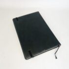 A closed black notebook with white stitching along the edges. An elastic band secures it, and a black ribbon bookmark extends slightly from the bottom edge. The background is a plain white surface.