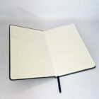 Open notebook with a black cover, displaying blank cream-colored pages. A black ribbon bookmark is visible in the middle. The notebook is on a plain white surface.